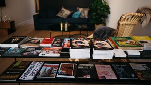 Una mesa con montones de revistas y libros, un sofá negro con cojines estampados, y una silla de mimbre en una sala de estar.