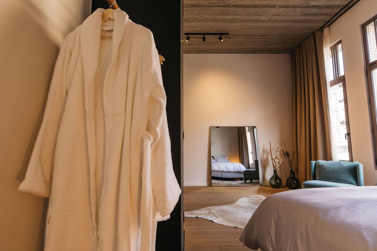 A hotel room with a large bed, a sitting area with a chair, a robe hanging on the wall, and a large mirror reflecting the room.