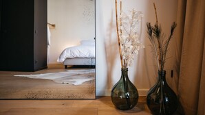 Una habitación con una cama, una puerta de vidrio que da a otra habitación, y dos jarrones con flores secas sobre un suelo de madera.
