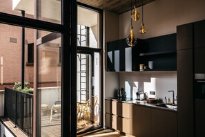 A modern kitchen with a large window, a dining area with wicker chairs, and a view of a brick building. - Charlie's B&B (Antwerp)