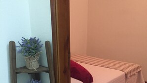 Un dormitorio con una estantería de madera con escalera que alberga plantas de lavanda, una cama con ropa blanca y un plaid rojo, y una pared blanca.