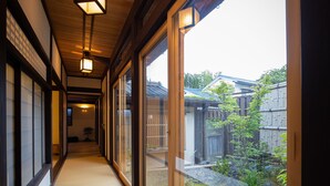 A corridor with tatami mat flooring, wooden beams, and sliding doors leading to a garden with a bamboo fence and stone pathway. - UCHIKO-Inn ORI (Uchiko)