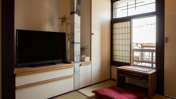 A traditional Japanese room with tatami flooring, a television on a wooden stand, a sliding door with a grid pattern, and a small table with a cushion.