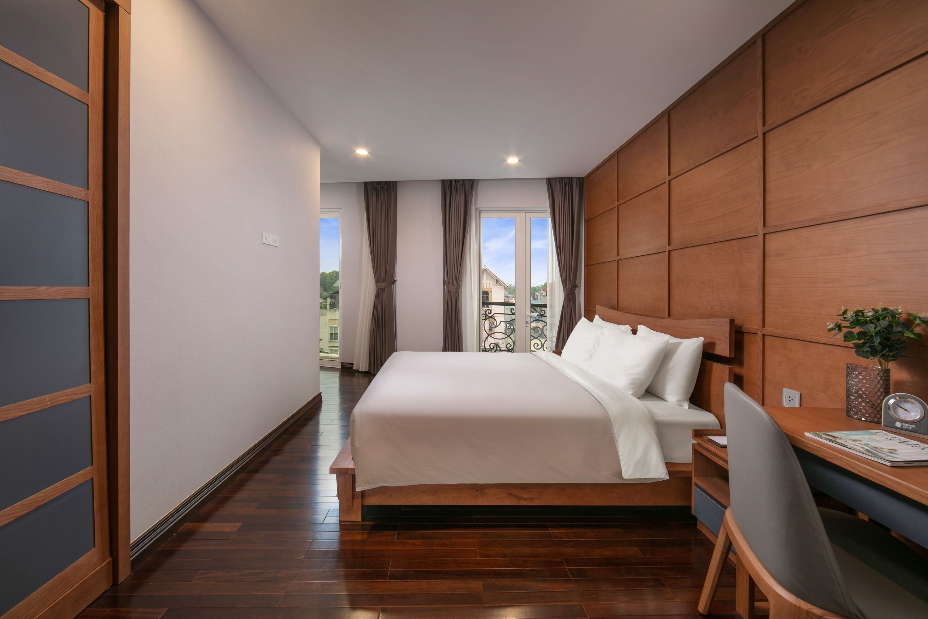 A modern bedroom with a wooden headboard, a large bed with white bedding, a desk with a chair, and a view of the outdoors through a window.