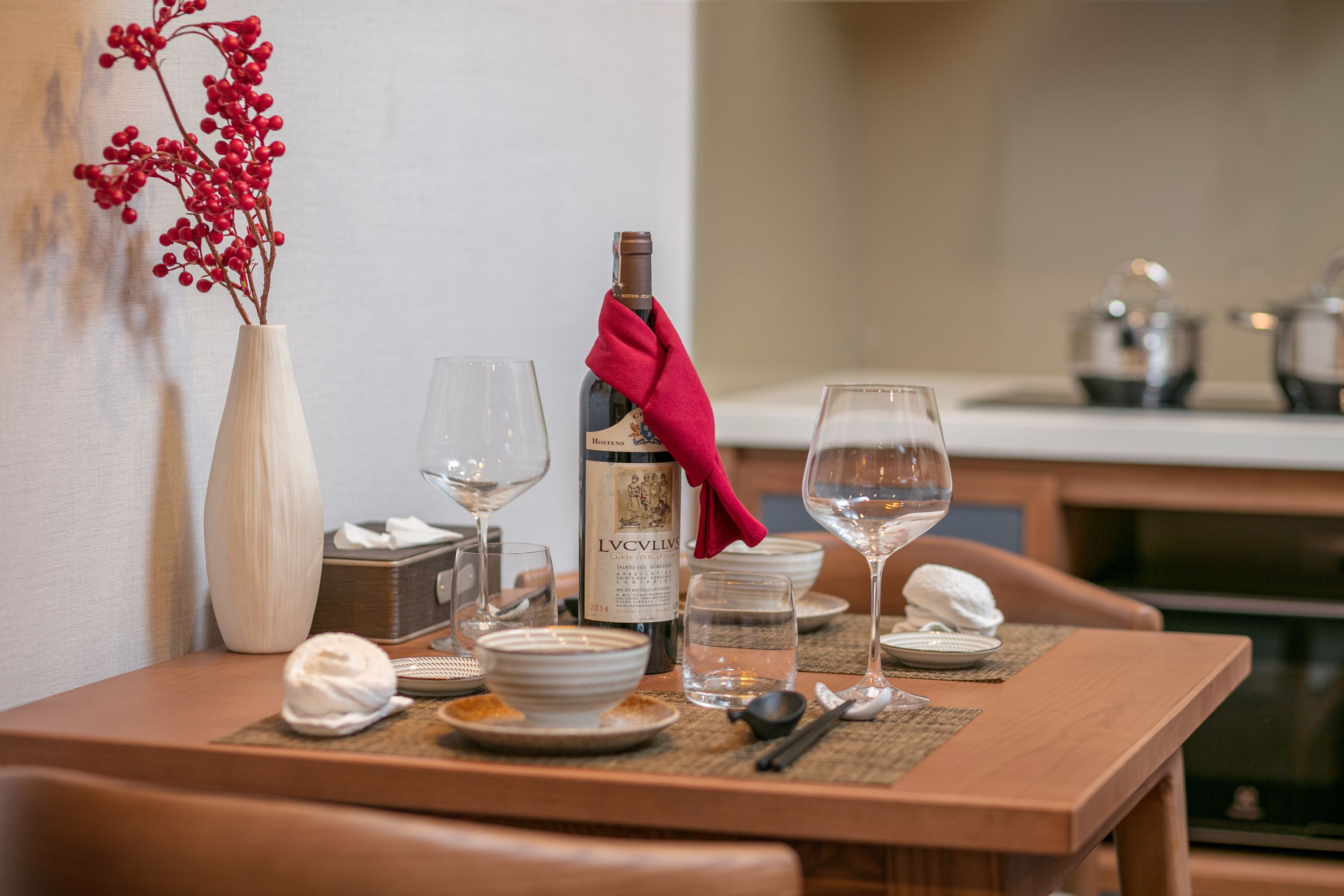 A dining table set with plates, bowls, glasses, and a bottle of wine, with a vase of red berries and a red napkin on the table.
