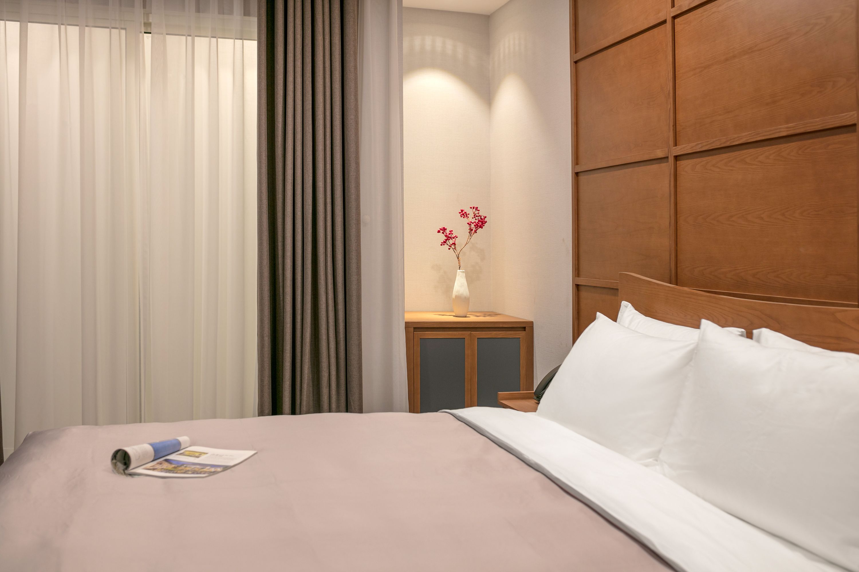 A neatly made bed with white linens, a wooden nightstand with a vase of flowers, and a window with sheer curtains.