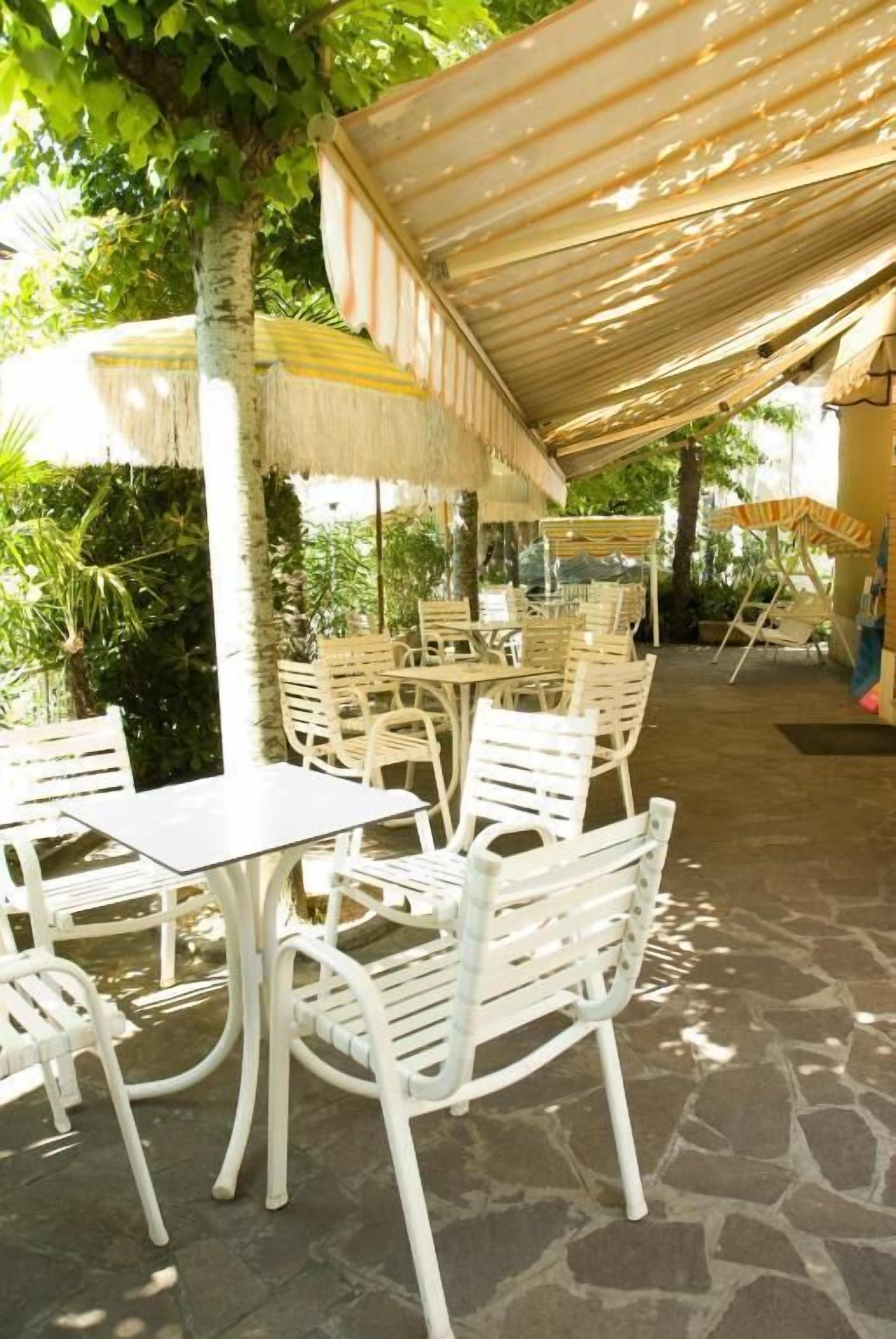 An outdoor patio with white metal chairs and tables, covered by striped awnings, surrounded by greenery.