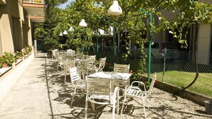 Un patio exterior con mesas y sillas, rodeado de follaje y una cerca.