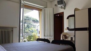 Habitación de hotel con cama, escritorio, silla, televisor y ventana con vista a árboles y edificios.