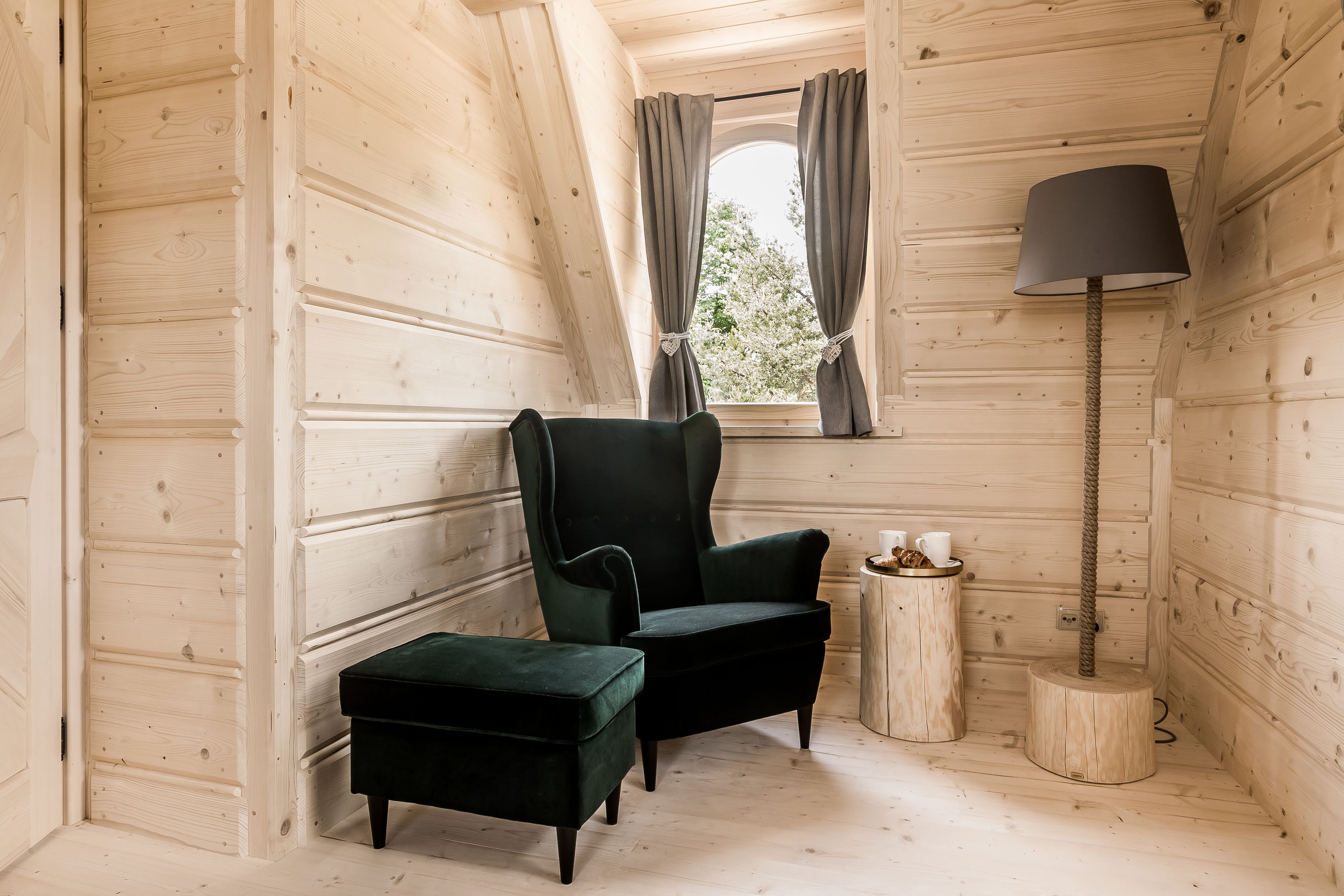 A cozy room with wooden walls, a dark green armchair, a matching ottoman, a small wooden side table with a lamp, and a window with curtains.