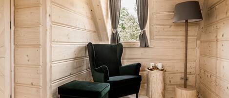 A cozy room with wooden walls, a dark green armchair, a matching ottoman, a small wooden side table with a lamp, and a window with curtains.