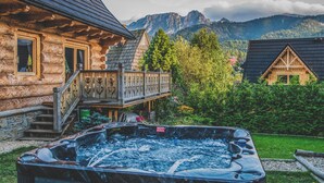 A wooden cabin with a hot tub, a balcony, and a mountainous backdrop.