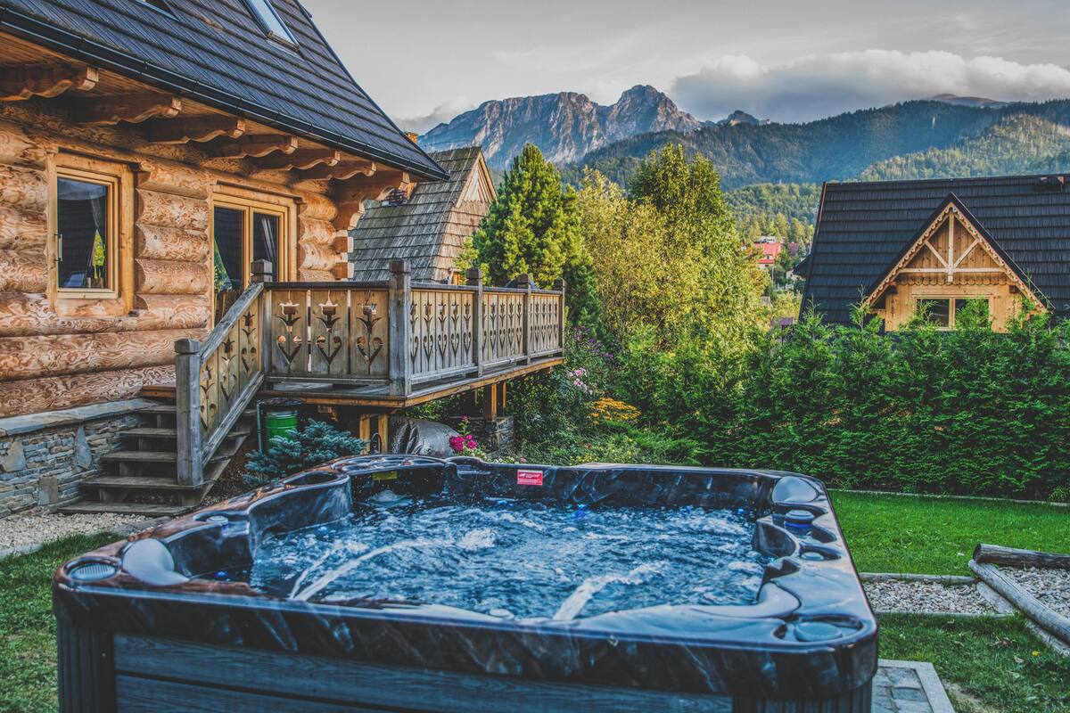 A wooden cabin with a hot tub, a balcony, and a mountainous backdrop.