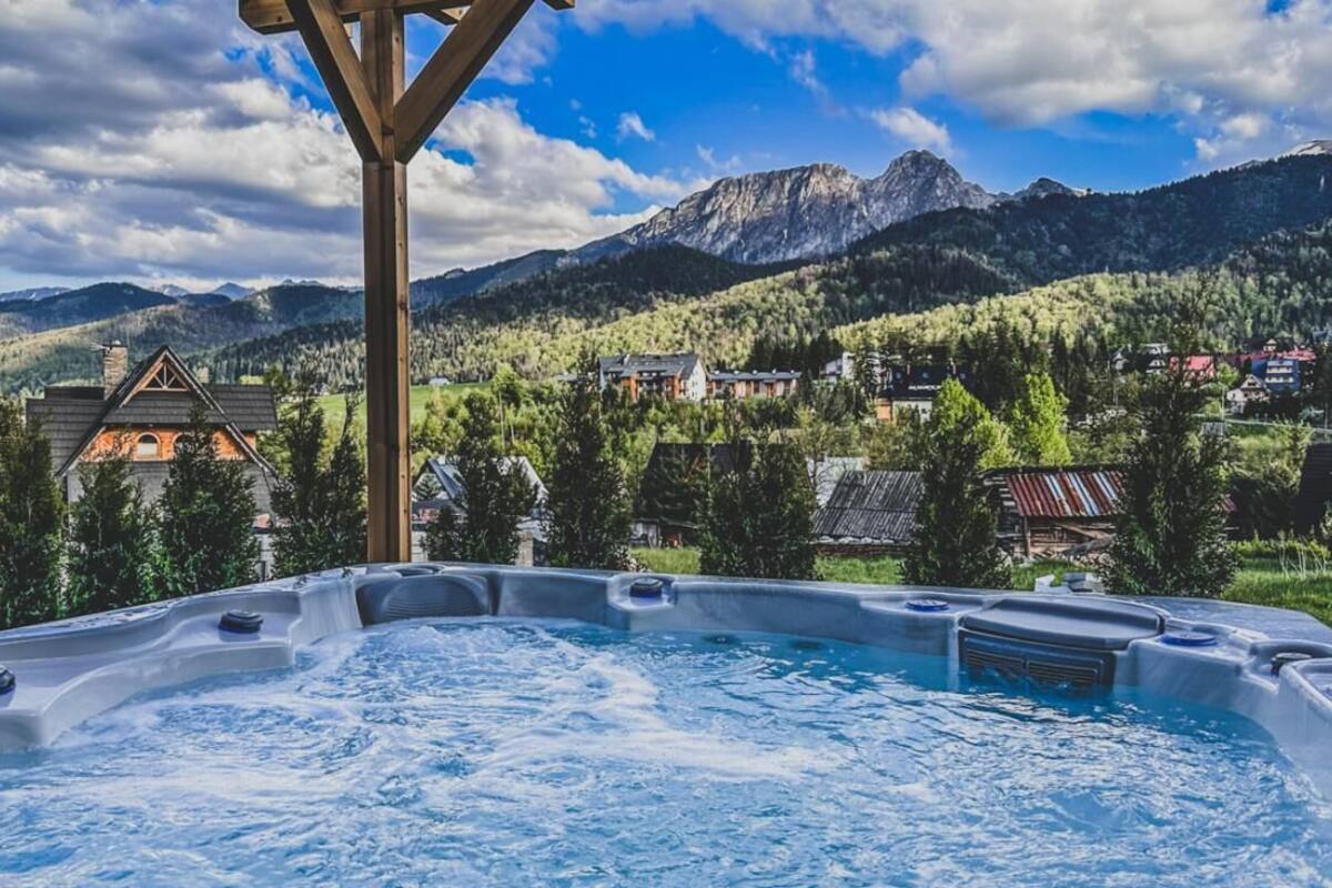 A hot tub with a wooden pergola structure, surrounded by a mountainous landscape and a village.