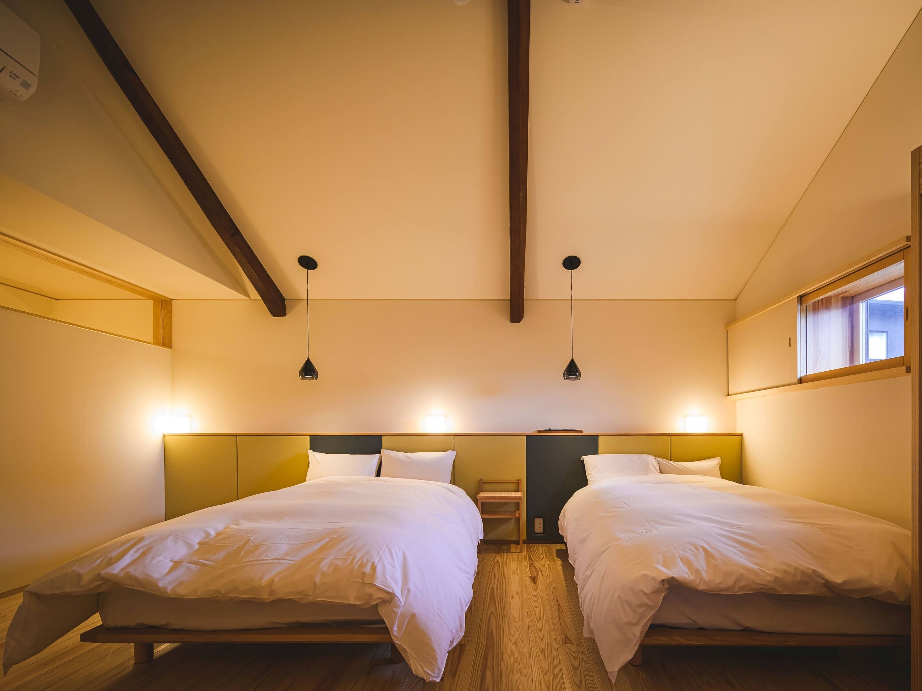 A hotel room with two beds, wooden flooring, and pendant lighting.