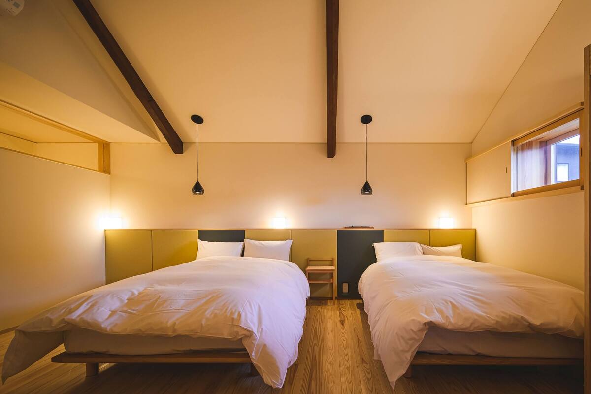 A hotel room with two beds, wooden flooring, and pendant lighting.