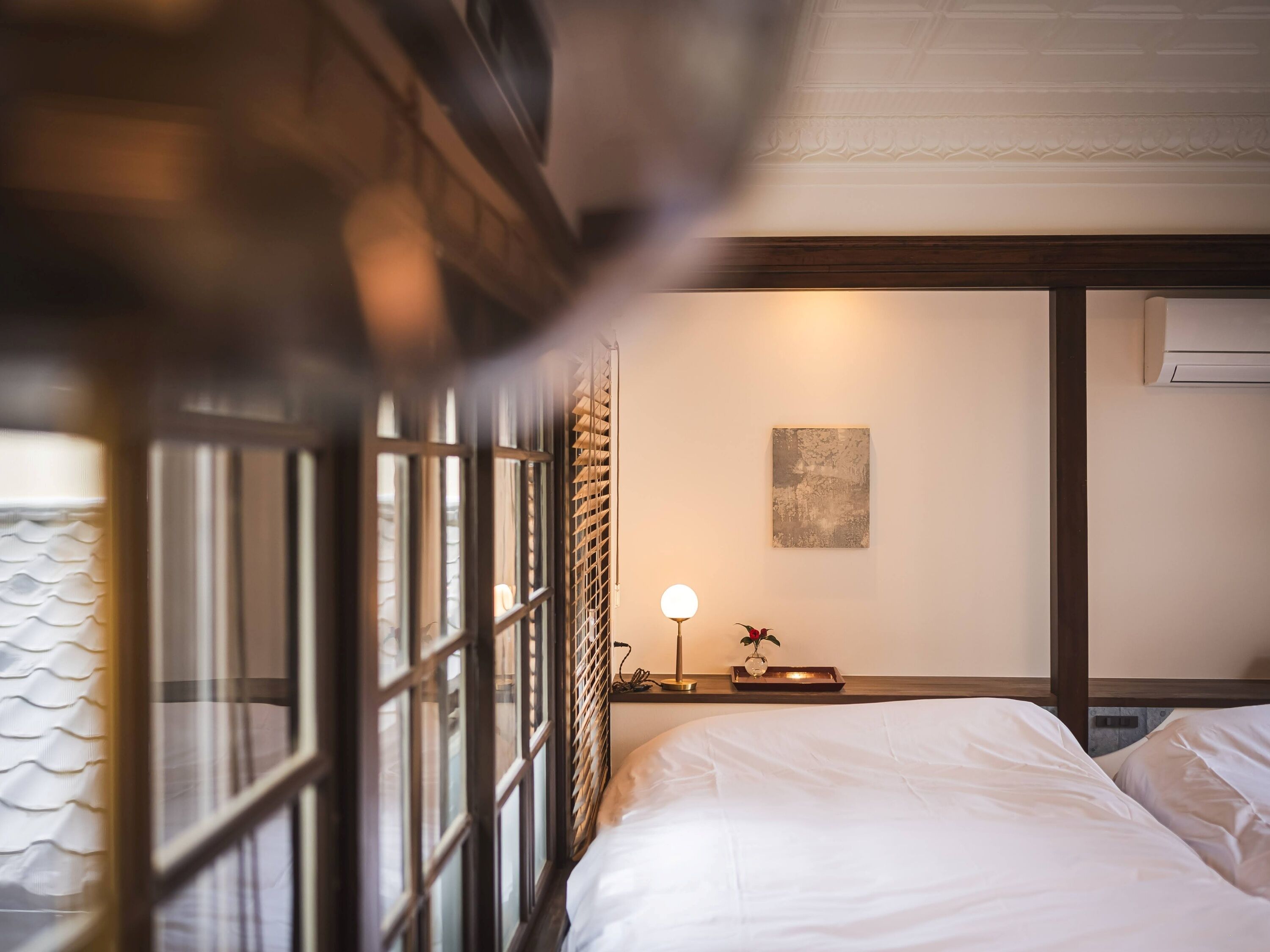 A hotel room with a bed, a bedside table with a lamp, a wall-mounted air conditioner, and a window with blinds.