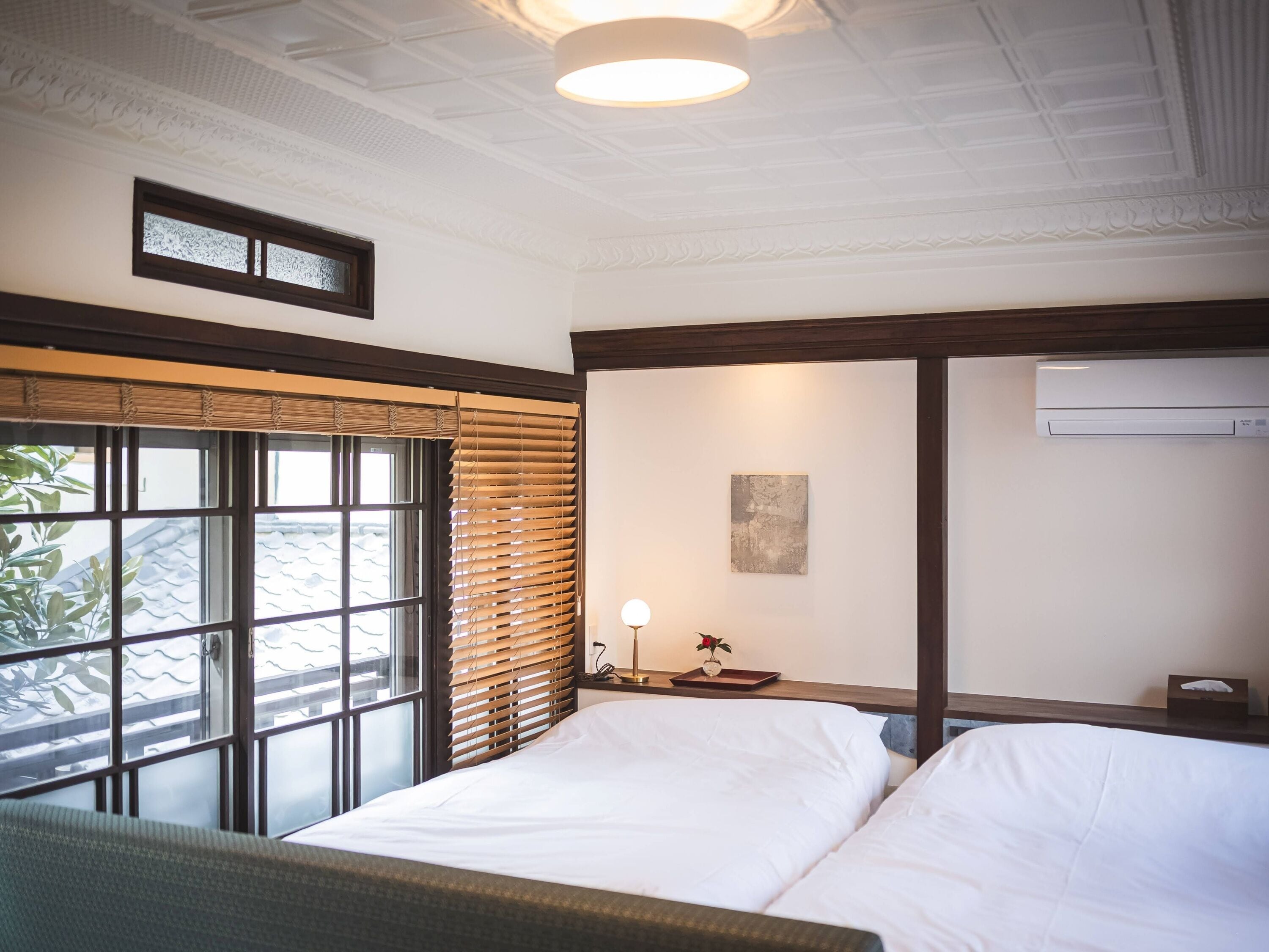 A hotel room with a large window, a bed, a bedside table with a lamp, and an air conditioning unit.