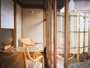 Une piĂšce avec une table en verre et des chaises en osier, un plancher en bois et une clĂŽture en bambou Ă lâextĂ©rieur.