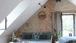 A loft-style bedroom with a sloped ceiling, exposed brick walls, a wooden floor, a bed, a sofa, a table, and a hanging light fixture. - Le Champ Bagnolet (Voussac)