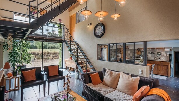 A spacious living area with a modern staircase, a large window offering a view of the outdoors, and a kitchen visible through the glass wall. - Le Champ Bagnolet (Voussac)