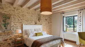 A bedroom with a stone wall, a bed with a wooden headboard, a hanging light fixture, and a window with curtains. - Le Champ Bagnolet (Voussac)