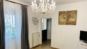 Un'ampia stanza con pavimento in legno, una grande finestra con tende, un lampadario, una mappa appesa al muro e un televisore su un mobile bianco.