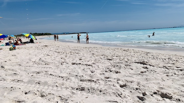 Una spiaggia con persone che passeggiano e nuotano, ombrelloni e un molo sullo sfondo.