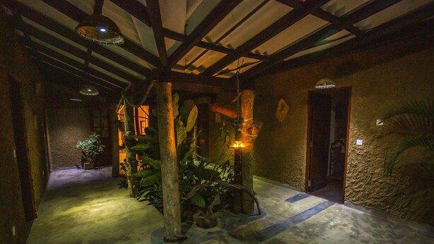 Un couloir faiblement éclairé, avec des poutres en bois rustiques et un sol en béton. On y aperçoit des plantes en pot et une petite porte.
