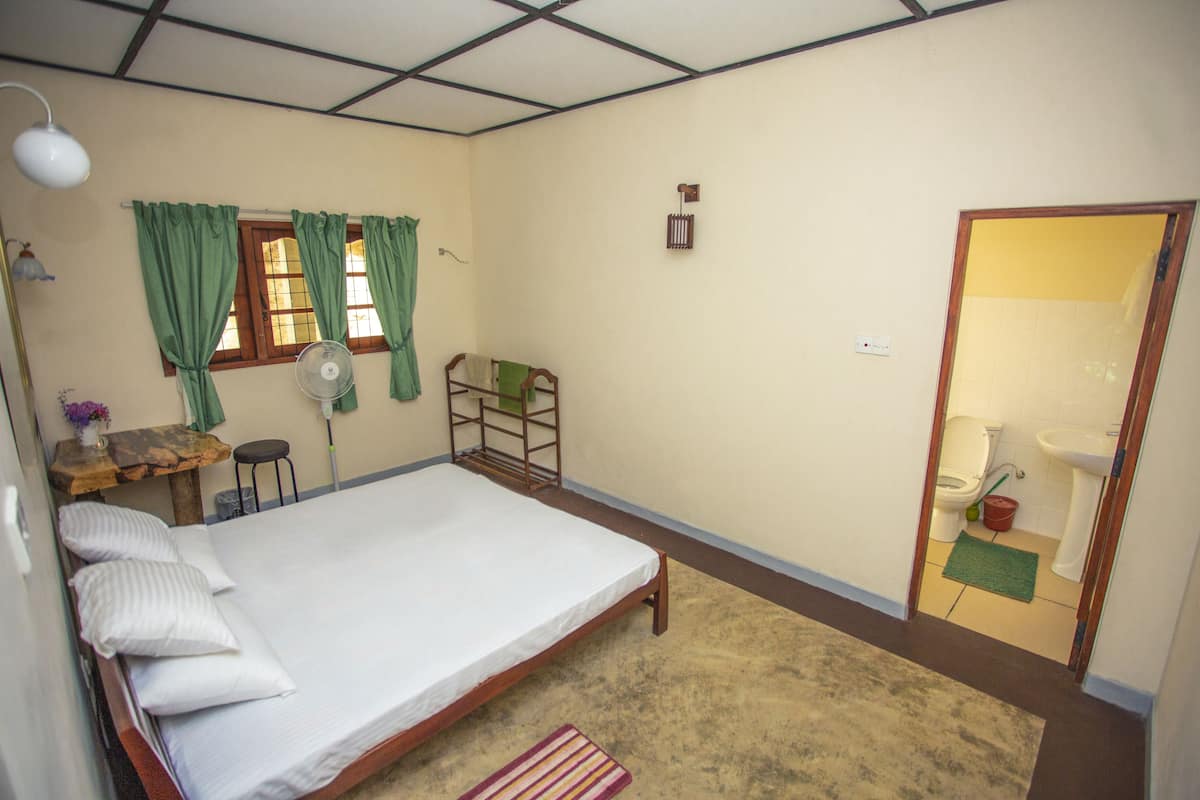 Ein Zimmer mit einem Einzelbett, einem Holzbett, einem kleinen Tisch mit Stuhl, einem Standventilator und einem Fenster mit grünen Vorhängen.