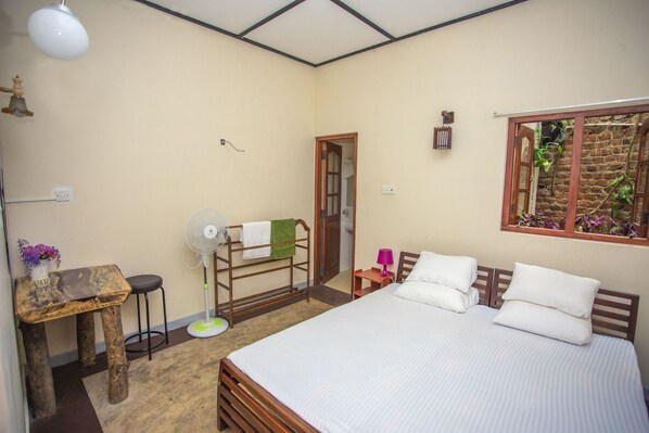 A bedroom with a bed, a small table, a fan, and a window with a view of plants. - The Hilltop Eco Homestay (Kundasale)