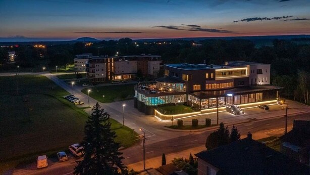 A modern building with a glass facade and a covered outdoor area, surrounded by a parking lot and greenery. - Hotel Kristály (Ajka)