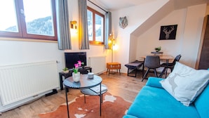 Ein gemütliches Wohnzimmer mit einem blauen Sofa, einem kleinen runden Tisch, einem Kamin und Blick ins Freie durch ein Fenster.