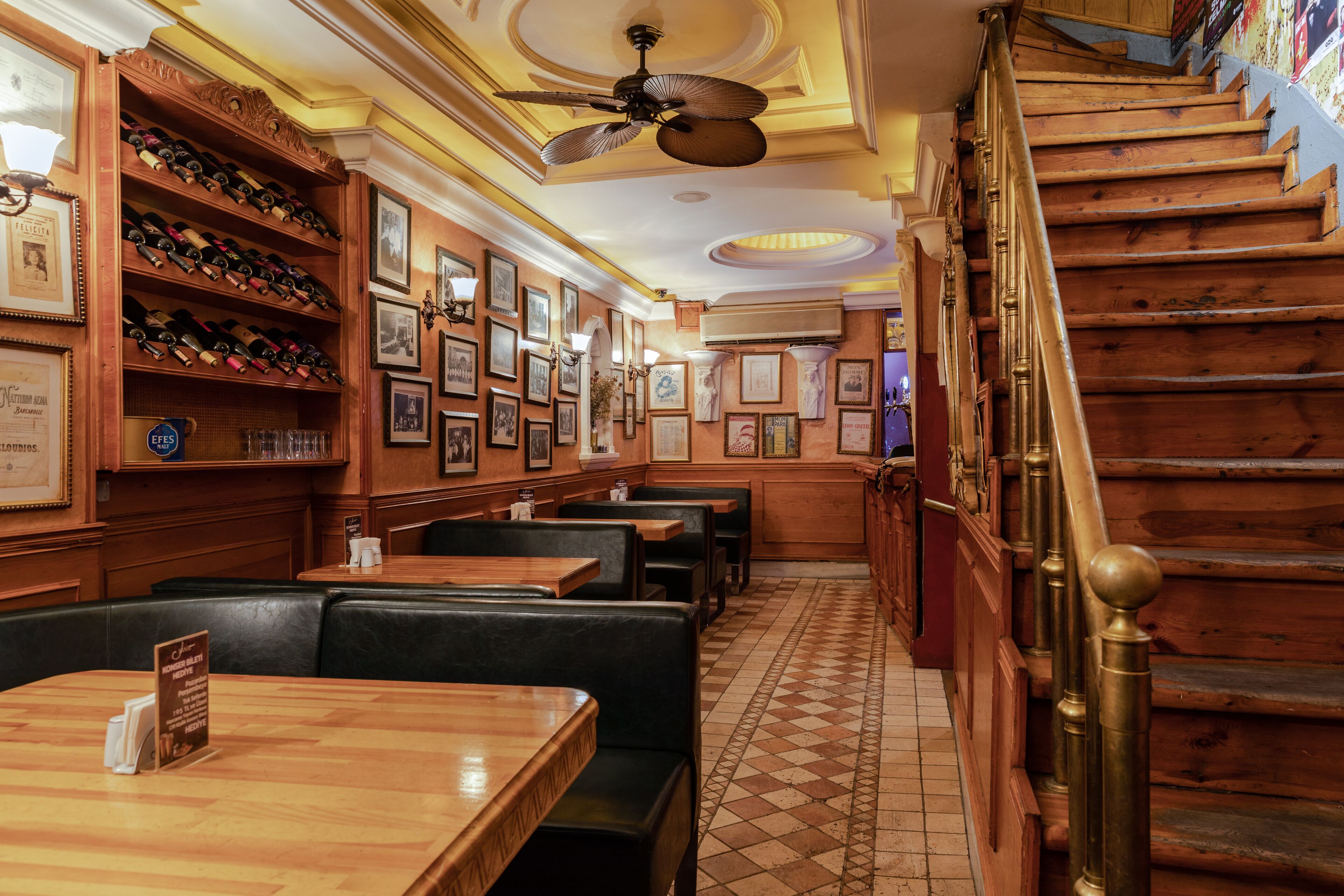 A restaurant interior with wooden tables and booths, a staircase with brass railings, and a ceiling fan.