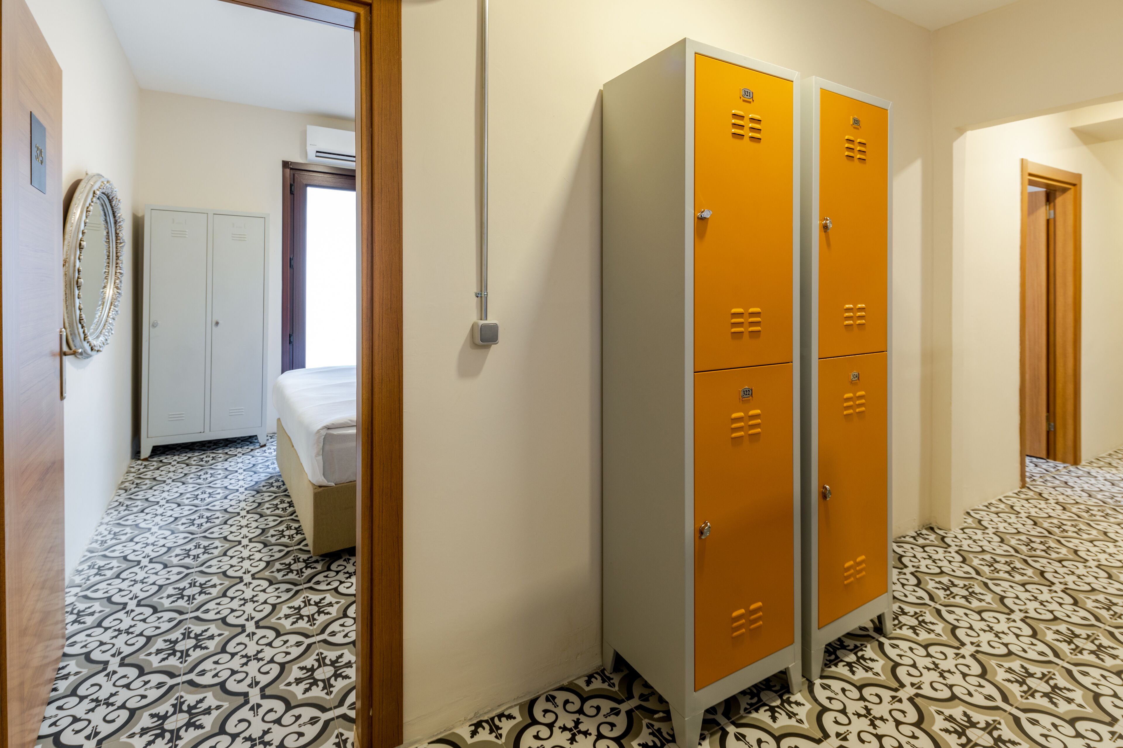 A hallway with patterned floor tiles, a bed, white and orange lockers, and a mirror on the wall.