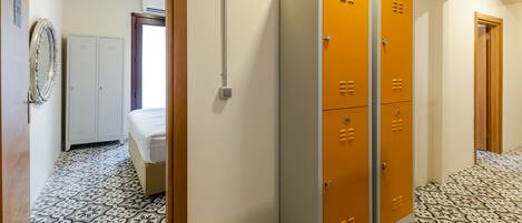 A hallway with patterned floor tiles, a bed, white and orange lockers, and a mirror on the wall.
