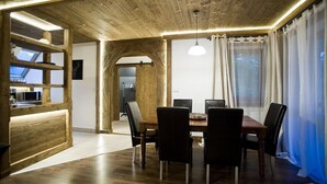 Een eethoek met een houten plafond, een centrale tafel met stoelen, en uitzicht op een keuken door een boog.