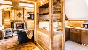 Interieur van een blokhut met een stenen openhaard, houten wanden, een keukenhoek en een zithoek met een stoel en een bank.