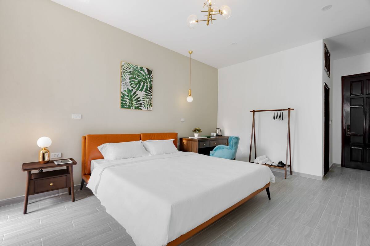 Una camera d'albergo moderna con un grande letto, un comodino con lampada, una scrivania con sedia, un appendiabiti e una porta.