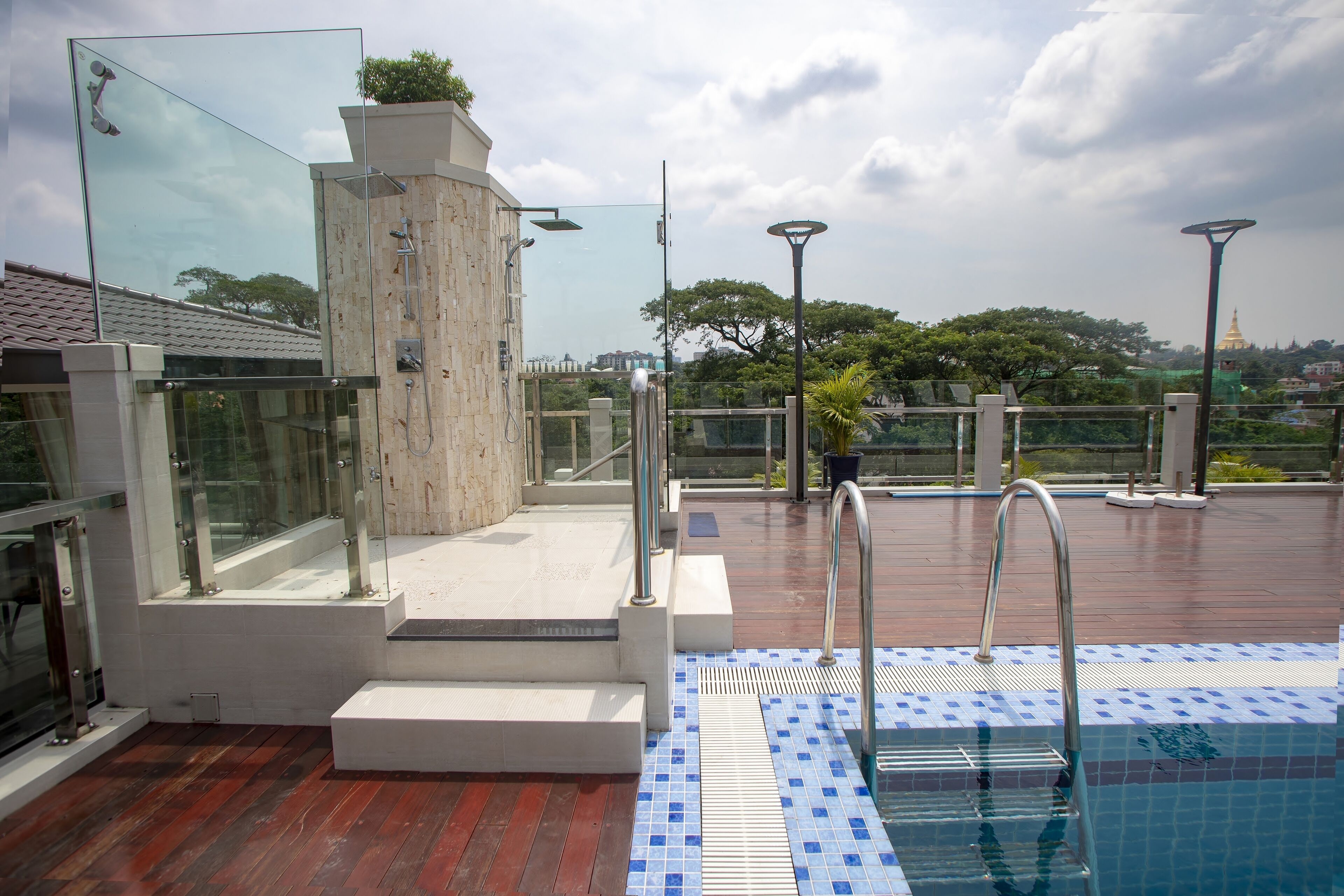 Un espace piscine aménagé sur le toit, doté de rampes en verre, d’une douche et offrant une vue sur un pagodon doré situé au loin.