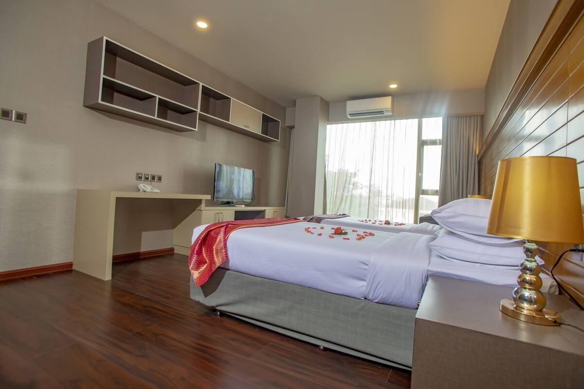 A hotel room with a bed, a desk with a television, a lamp, and a window with curtains.