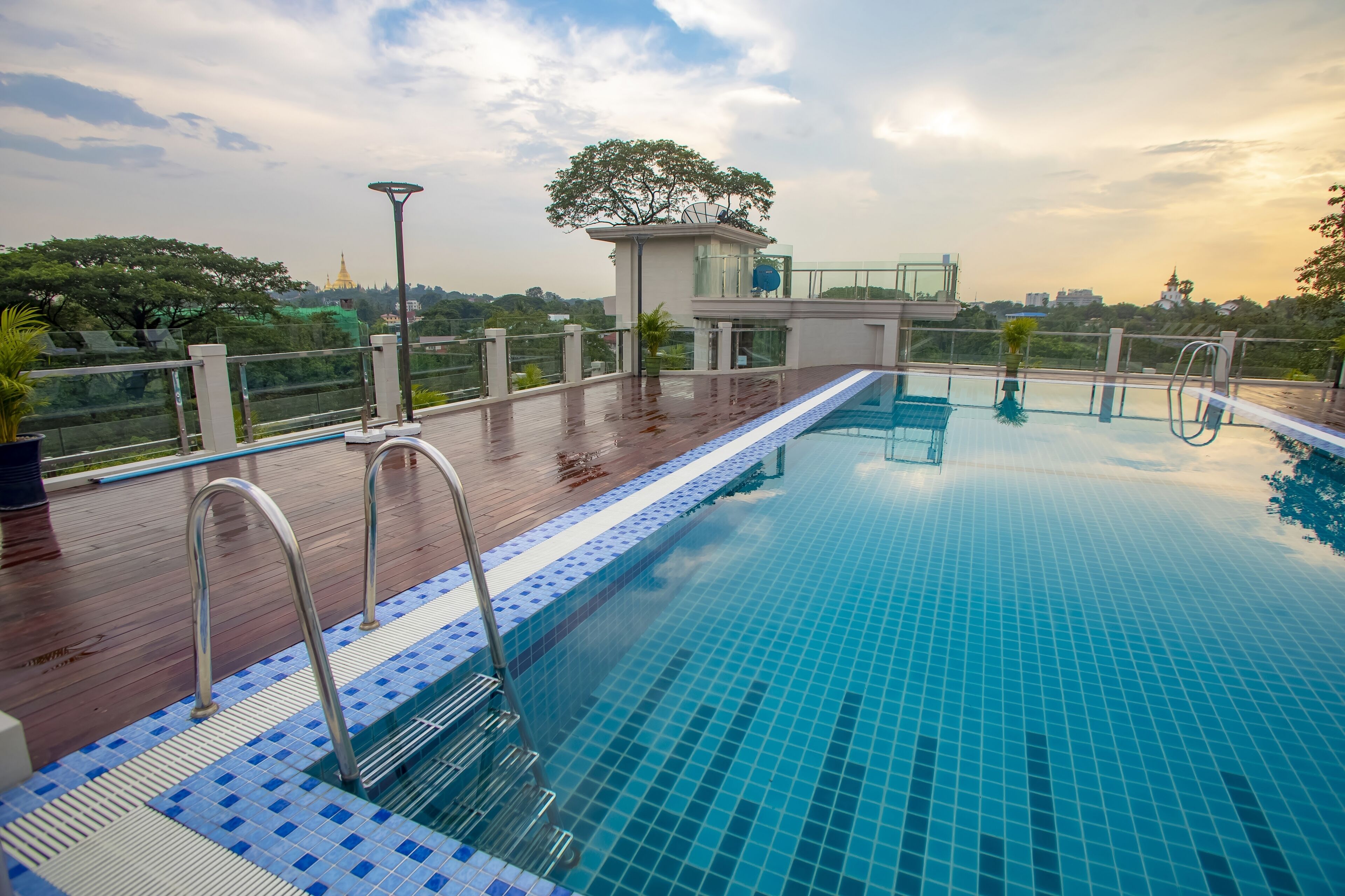 A rooftop swimming pool with a wooden deck, metal railings, and a view of a temple in the background.