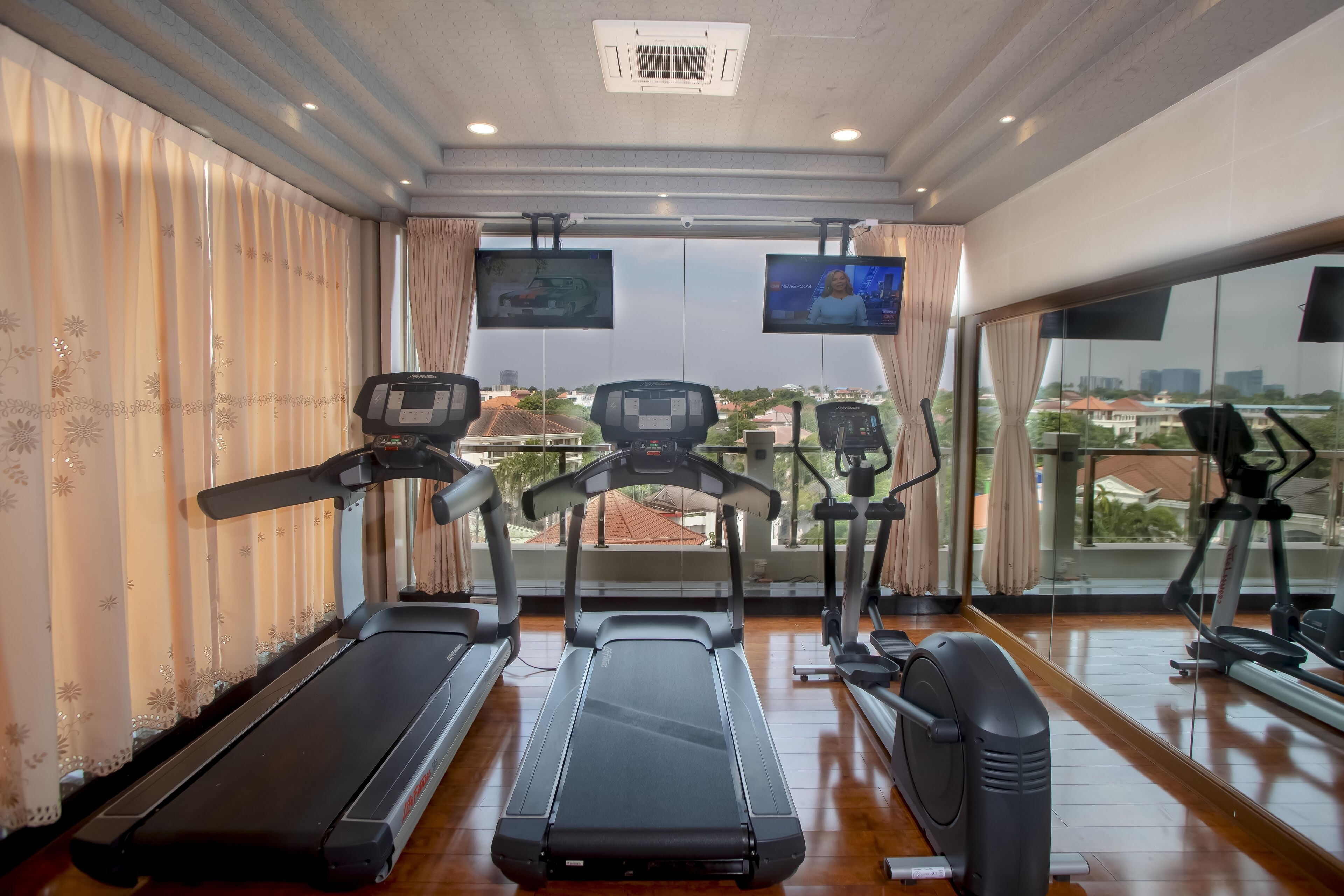 Un gimnasio con cintas de correr, una bicicleta estática y una máquina de remo. Hay dos televisores montados en la pared, y la habitación tiene ventanas grandes con vistas a edificios.