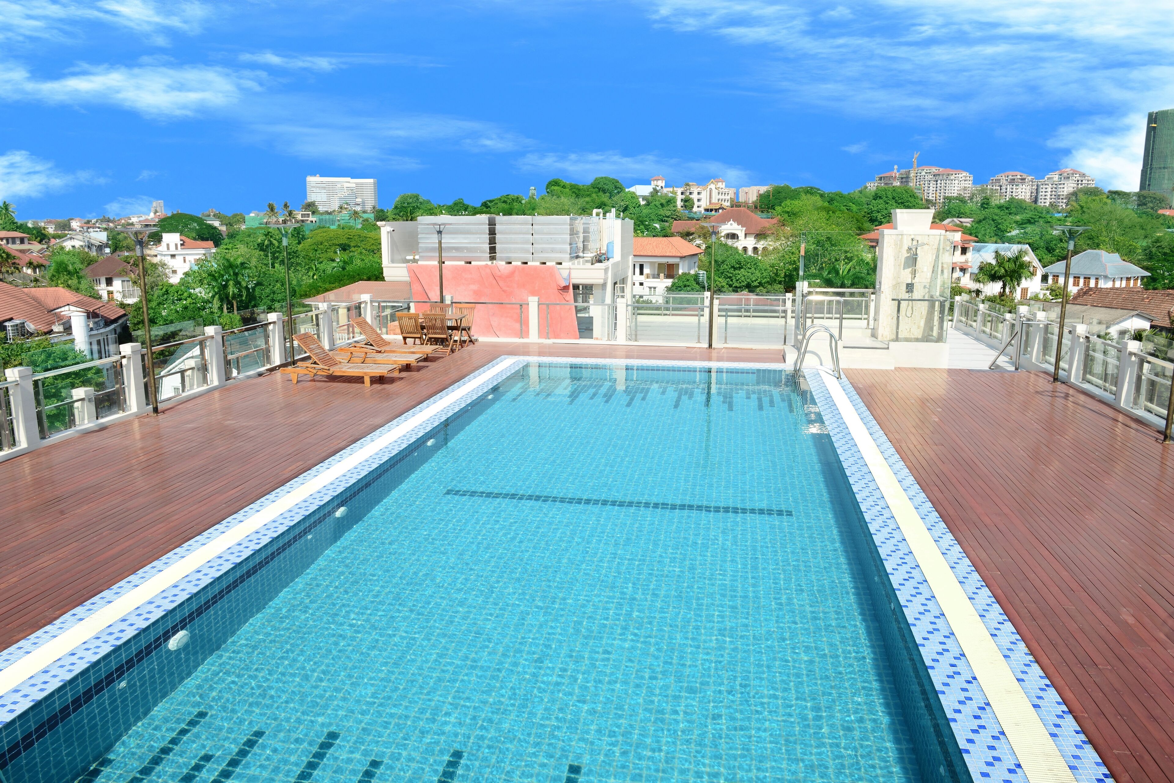 Una piscina en la azotea con terraza de madera, rodeada por una barandilla y vistas a un paisaje urbano con edificios y zonas verdes.