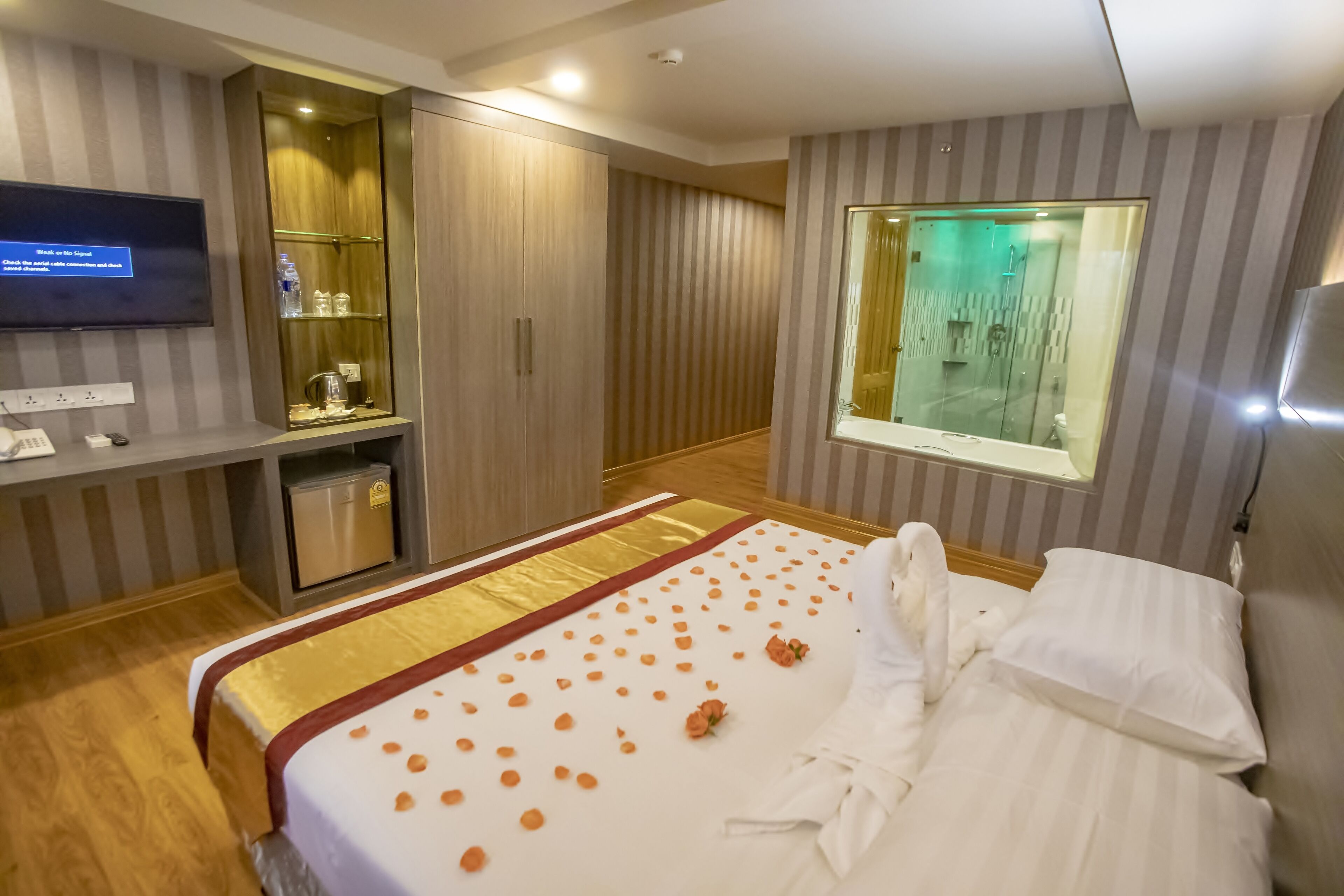 Habitación de hotel con cama, televisión, minibar y baño visible a través de una ventana.