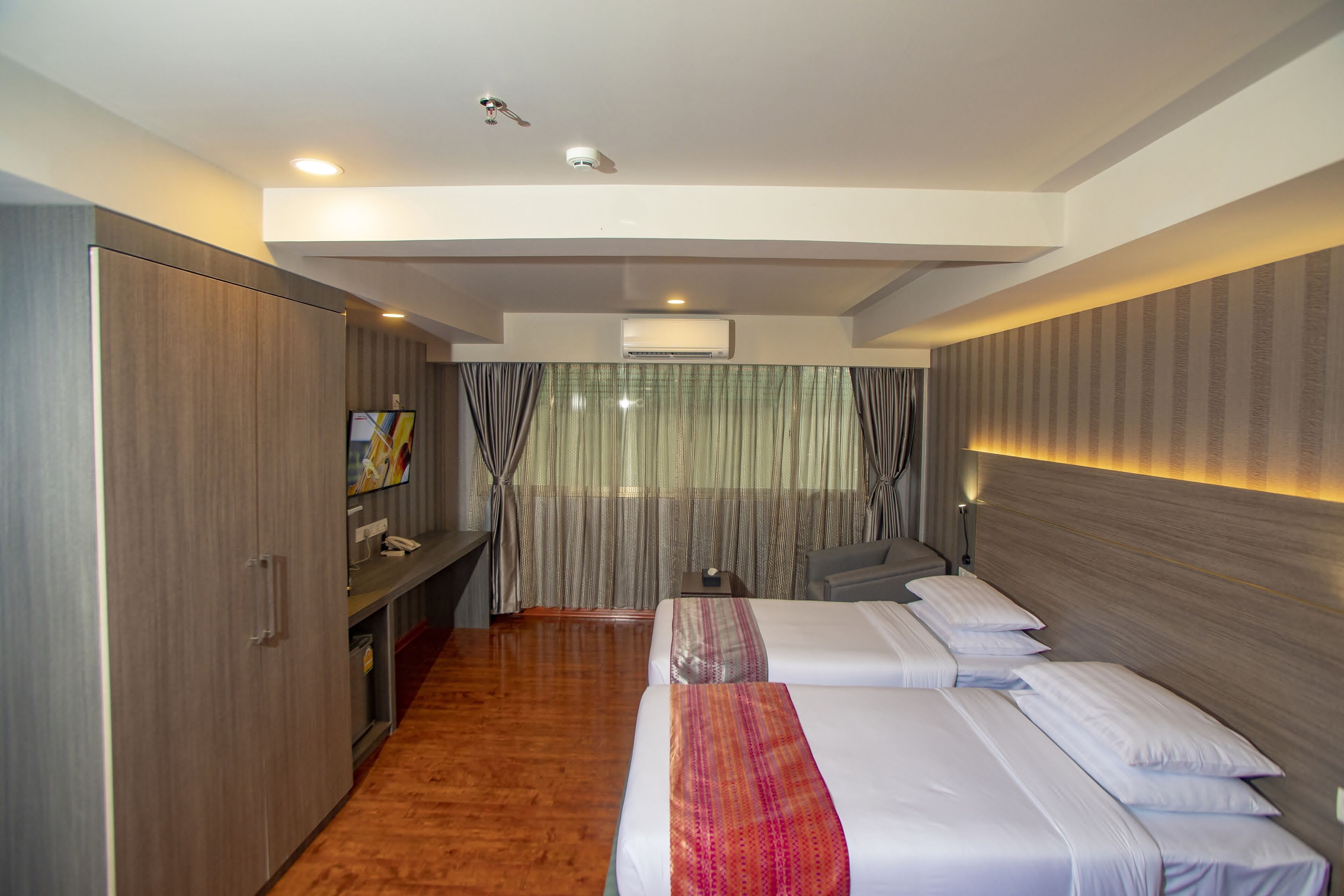 A hotel room with a wooden floor, a bed with white bedding, a desk with a chair, and a wardrobe.