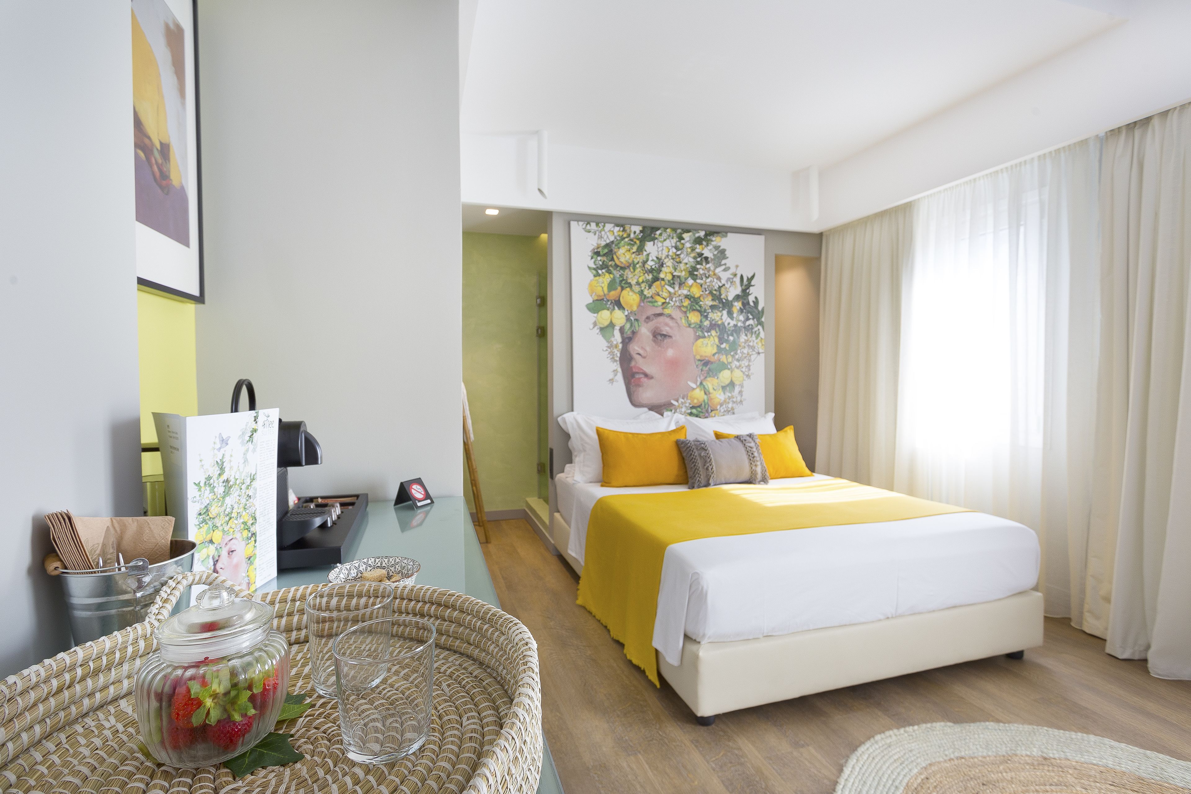 A hotel room with a bed, a bedside table, a wicker chair, a glass jar with strawberries, and a framed picture on the wall.