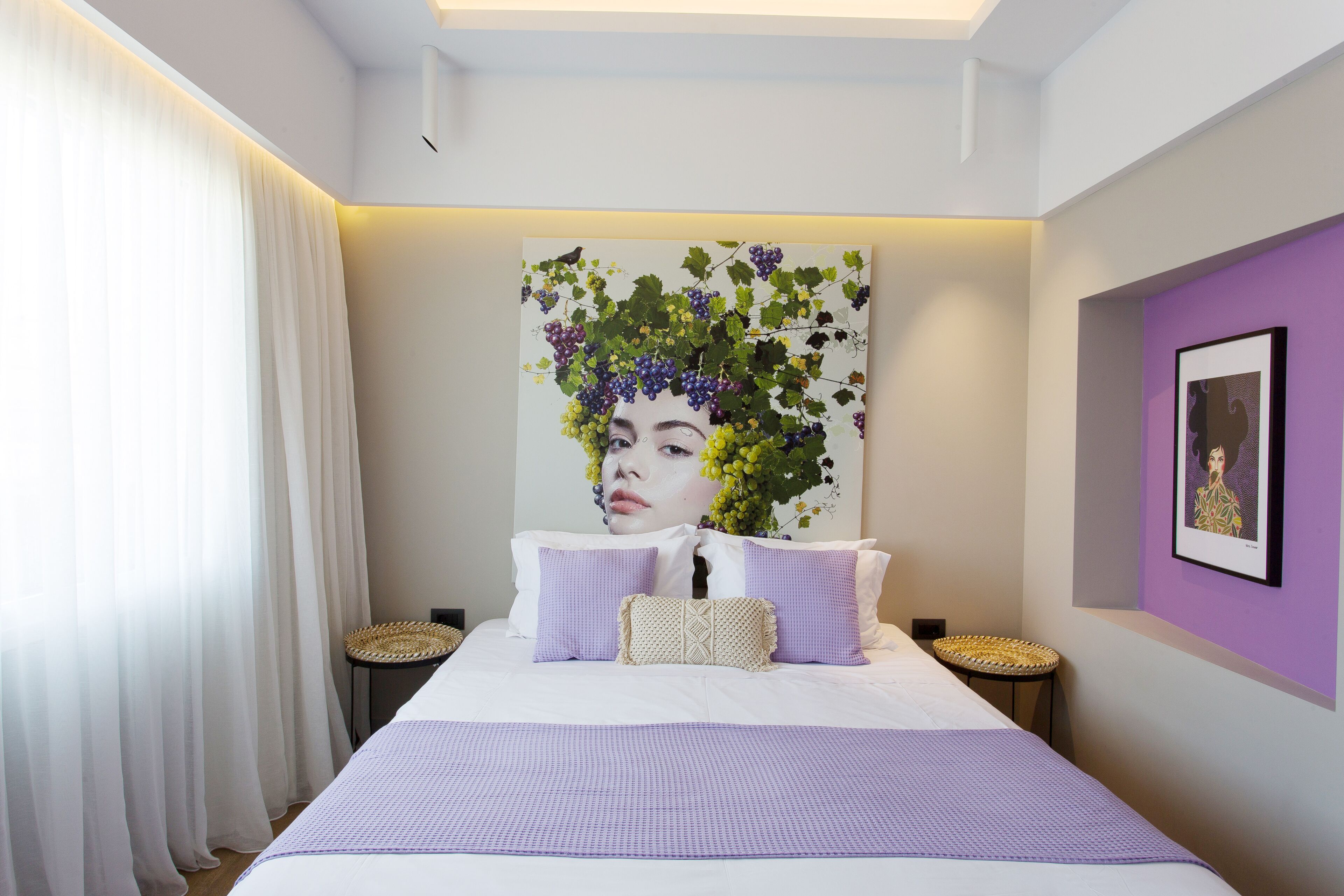Een slaapkamer met een groot bed, wit beddengoed, een kunstwerk aan de muur waarop een vrouw te zien is omringd door druiven, en een paarse accentmuur.
