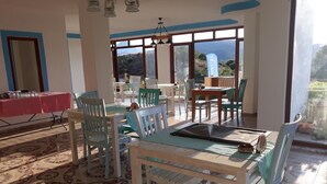 A dining area with a table set for two, surrounded by chairs, and a view of a mountainous landscape through large windows. - ADA KEKİĞİ PANSİYON (Gökçeada)