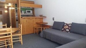 Kompakti huone, jossa on puinen ruokapöytä ja -tuolit, harmaa sohva kuviollisilla koristetyynyillä sekä pieni keittokomero, jossa on mikroaaltouuni ja pieni hylly.